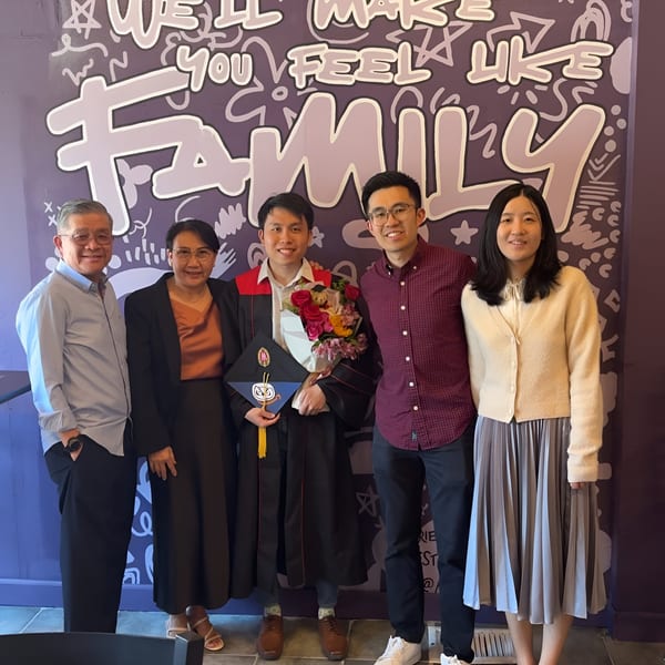 Five people, including a graduate, smiling in front of a purple wall with "WE'LL MAKE YOU FEEL LIKE FAMILY" written on it.