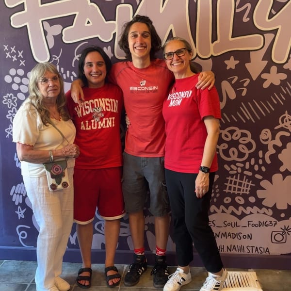 Four people, likely a family, smiling in front of a purple wall with "FAMILY" painted on it.